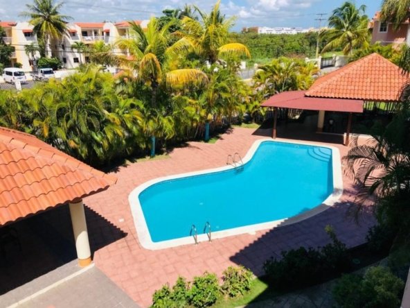 Resort pool area with blue water, red-tiled deck, and surrounding palm trees under a sunny sky